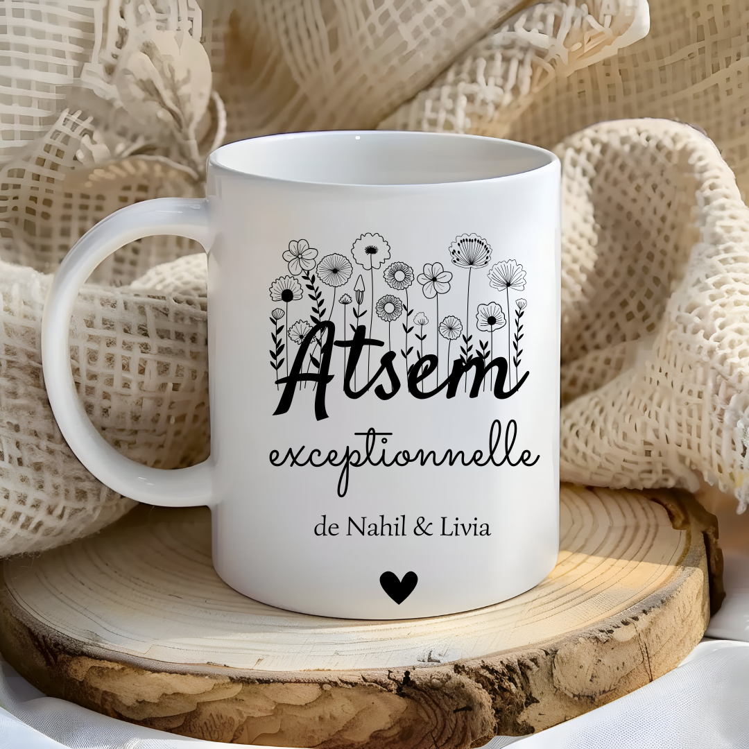  Mug blanc avec l’inscription "atsem Exceptionnelle" et le prénom "Lucas" posé sur une table entourée de fournitures scolaires.