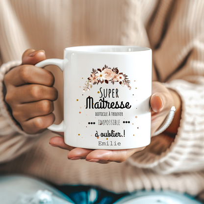  Mug blanc avec l’inscription "Super Maîtresse – Difficile à trouver, impossible à oublier" posé sur un bureau avec des fournitures scolaires.